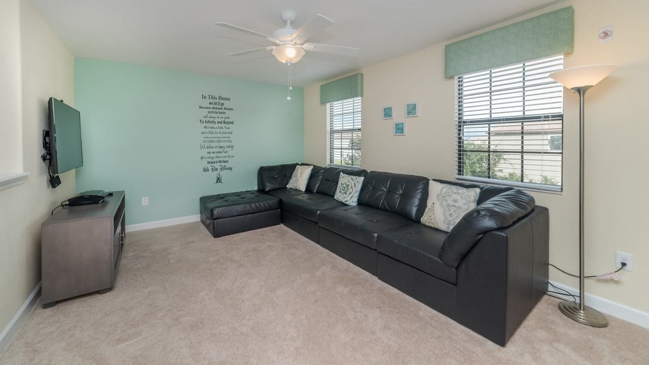 A living room with a sectional sofa, a ceiling fan, a flat-screen TV, a floor lamp, and a wall with a motivational quote.