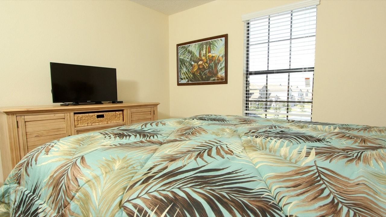 Un dormitorio con cama, televisor sobre una cómoda de madera, ventana con persianas y un cuadro en la pared.