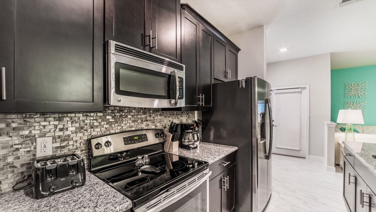 A modern kitchen with dark cabinets, a stainless steel microwave, a refrigerator, a stove, and a tiled backsplash.