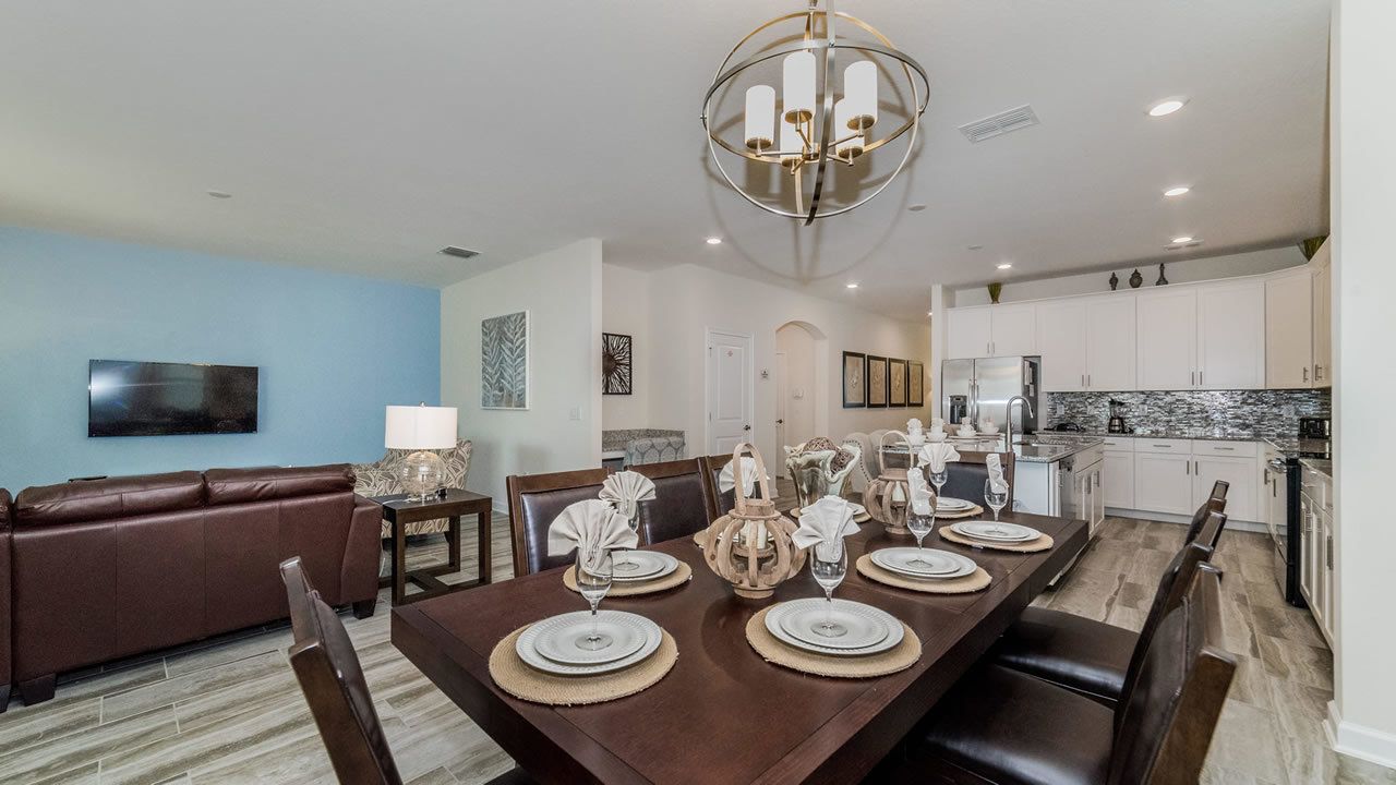 A dining area with a long table set for six, a brown leather sofa, and a kitchen in the background.