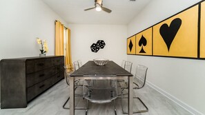 A dining area with a long table, chairs, a dark wooden cabinet, a decorative wall piece, and a ceiling fan. - Grhmvd1688 - Champions Gate Resort - 6 Bed 6 Baths House (Davenport)