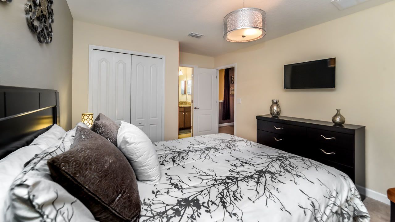 A bedroom with a bed, bedside tables, a dresser, a TV, and a door leading to another room.