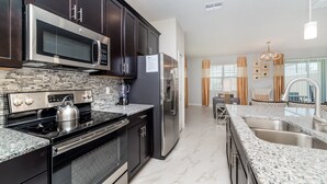 A modern kitchen with dark cabinets, stainless steel appliances, a granite countertop, and a view into the living area.
