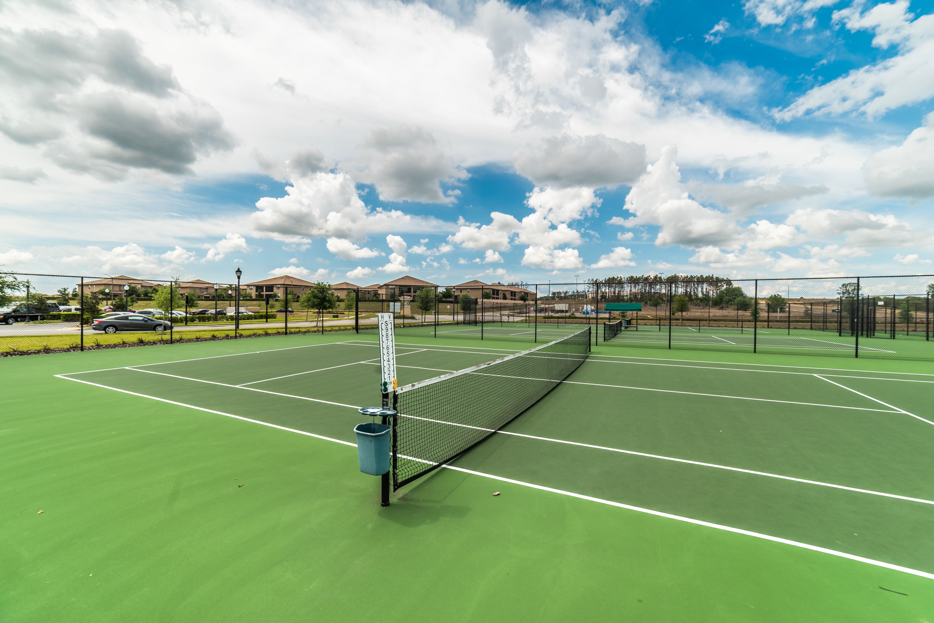 A tennis court with a net, surrounded by a fence, and houses in the background.