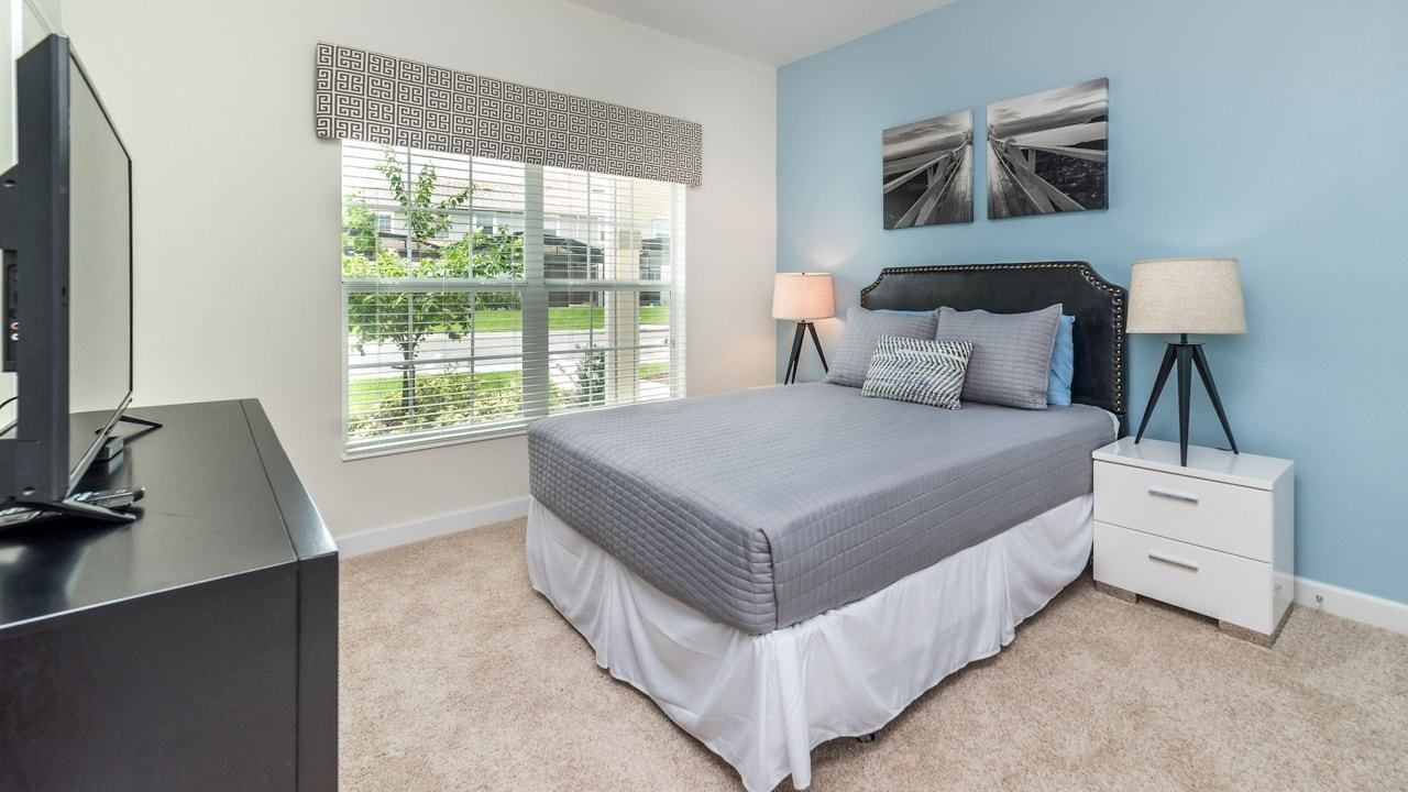 A bedroom with a bed, bedside tables, a TV, and a window with blinds.
