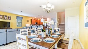 A dining area with a wooden table set for four, surrounded by white chairs. A chandelier hangs above the table, and there is a staircase leading to another level. - Grhcdw9151 - Champions Gate Resort - 9 Bed 5 Baths House (Davenport)