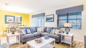 A living room with a grey sectional sofa, white coffee tables, and a dining area with a white table and chairs. - Grhcdw9151 - Champions Gate Resort - 9 Bed 5 Baths House (Davenport)