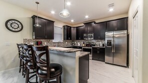 Una cocina moderna con armarios oscuros, electrodomésticos de acero inoxidable, una isla central con taburetes y un reloj en la pared.