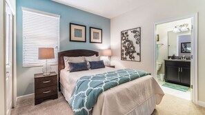 A bedroom with a bed, bedside tables, a lamp, a window with blinds, a framed artwork, and a bathroom visible through an open door. - Grhcdw9157 - Champions Gate Resort - 6 Bed 6 Baths House (Davenport)