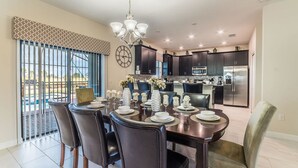 A dining area with a large table set for six, surrounded by dark chairs, a chandelier, and a view of the outdoor patio through large windows. - Grhcdw9157 - Champions Gate Resort - 6 Bed 6 Baths House (Davenport)