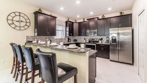 A modern kitchen with dark cabinets, stainless steel appliances, a granite countertop, and a clock on the wall. - Grhcdw9157 - Champions Gate Resort - 6 Bed 6 Baths House (Davenport)