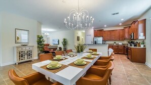 A spacious dining area with a long table set for six, surrounded by orange chairs. A modern chandelier hangs above, and the room features wooden cabinets and a tiled floor. - Grhwgd9165 - Champions Gate Resort - 4 Bed 3 Baths House (Davenport)