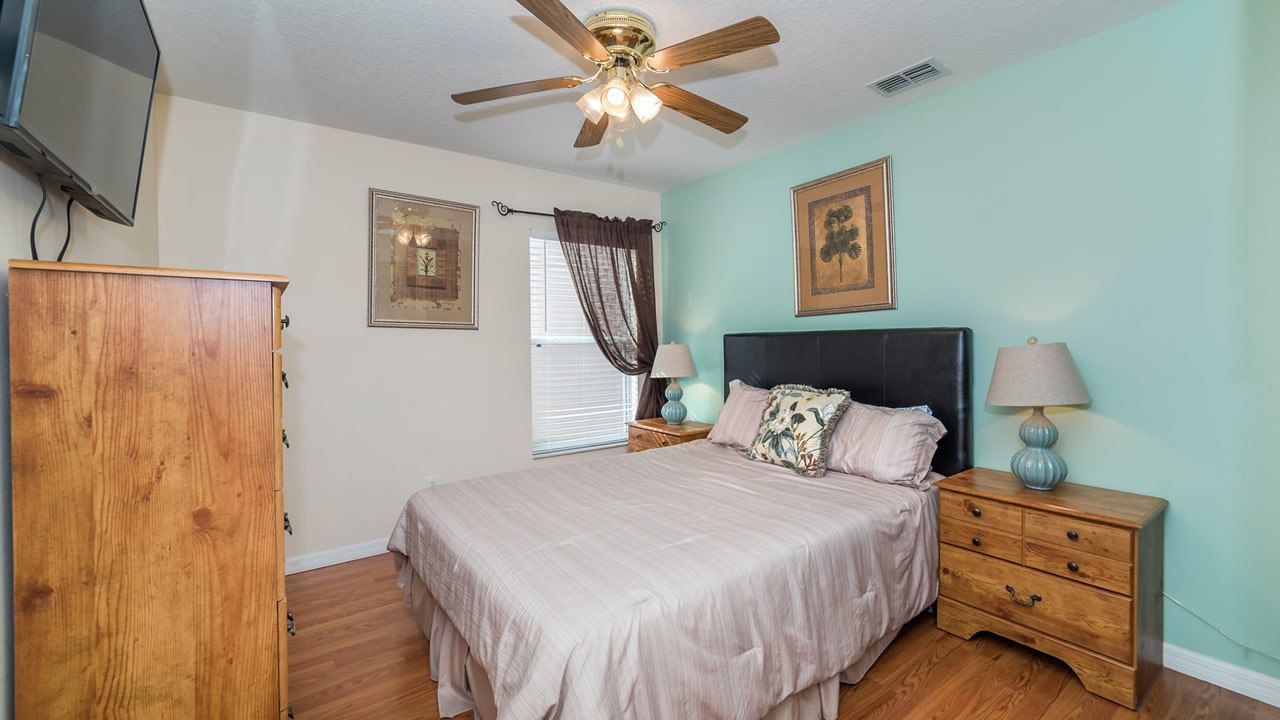 A bedroom with a bed, wooden nightstand, ceiling fan, and a TV mounted on the wall.