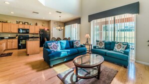 A spacious living room with a blue sofa set, a glass coffee table, and a kitchen area with wooden cabinets and a black refrigerator.