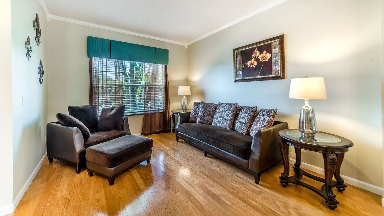 Un salon avec un canapé marron foncé, un pouf assorti, deux tables d’appoint avec des lampes, une fenêtre avec des stores et un tableau encadré au mur.