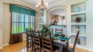 A dining area with a table set for four, wooden chairs, a chandelier, and a view of a green outdoor space through large windows.