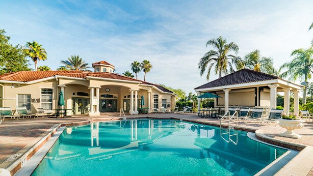 A swimming pool with lounge chairs and umbrellas, surrounded by palm trees and a building with a tiled roof. - Grhokc8484 - Emerald Island Resort - 5 Bed 4 Baths House (Kissimmee)
