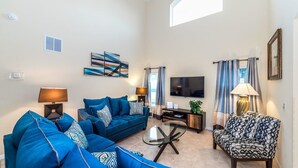 A living room with a blue sofa, a patterned armchair, a glass coffee table, and a flat-screen TV. - Grhokc8484 - Emerald Island Resort - 5 Bed 4 Baths House (Kissimmee)