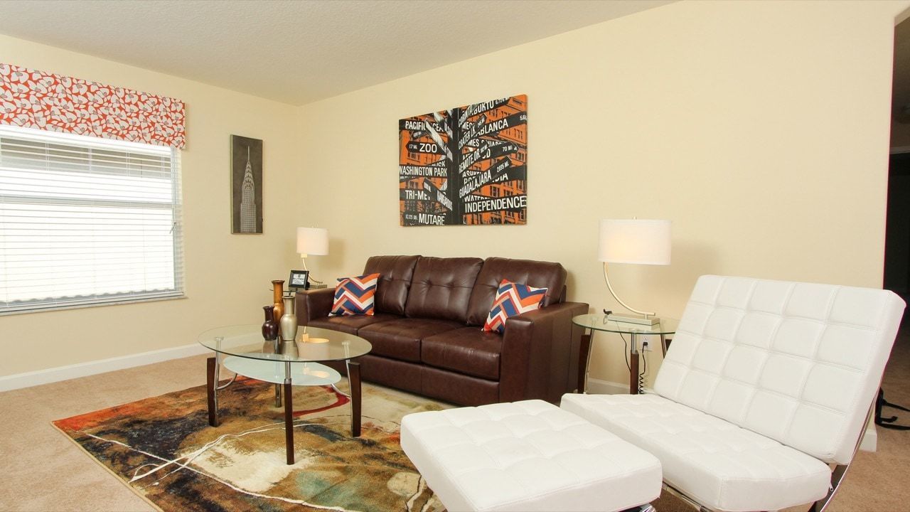 A living room with a brown leather sofa, a white armchair, a glass coffee table, and a patterned rug. There is a wall art piece above the sofa and a lamp on a side table.