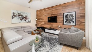 A modern living room with a sectional sofa, a grey armchair, a coffee table with a plant, a flat-screen TV mounted on a wooden wall, and abstract art on the walls. - Grhbch2953 - Paradise Palms Resort - 6 Bed 5 Baths House (Kissimmee)