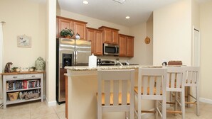 A kitchen with wooden cabinets, a stainless steel refrigerator, a microwave, and a dining area with white chairs. - Grhbch3057 - Paradise Palms Resort - 5 Bed 4 Baths Townhouse (Kissimmee)