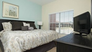 Une chambre à coucher avec un lit, un téléviseur posé sur une commode, une fenêtre avec des stores et un tableau accroché au mur.