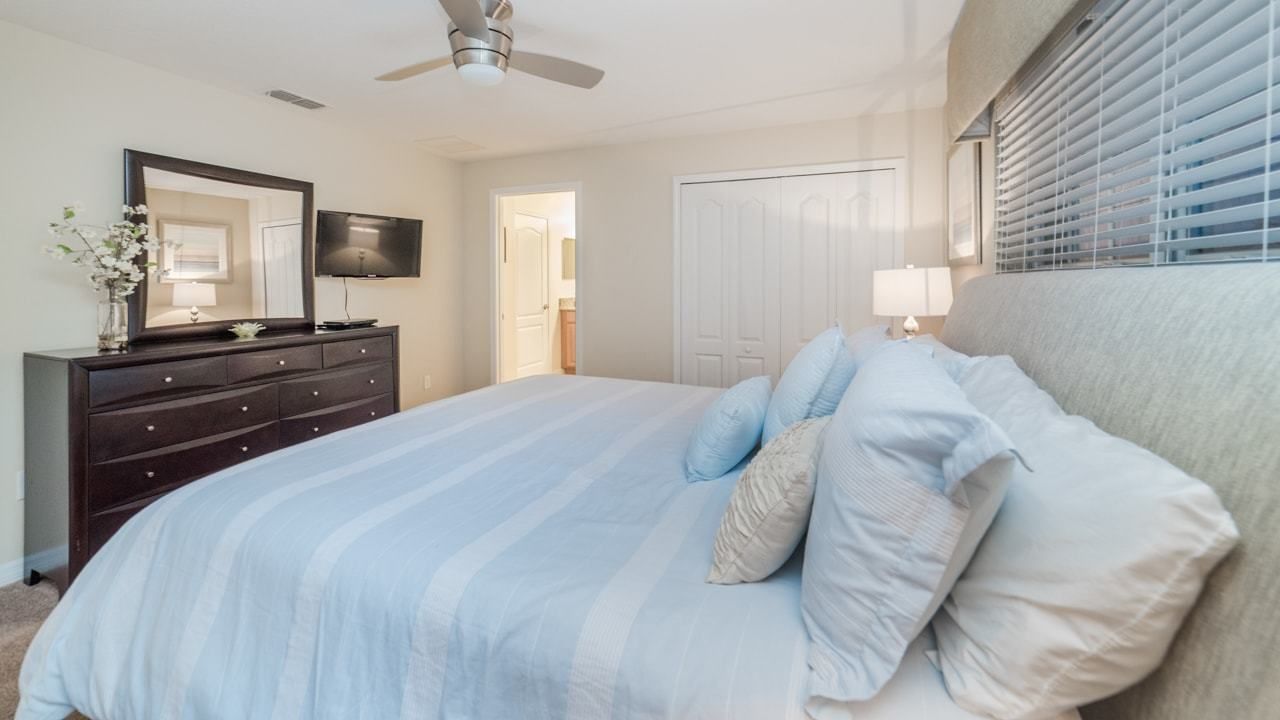 A bedroom with a bed, dresser, TV, ceiling fan, and a door leading to another room.