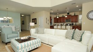 A modern living room with a white sofa, a patterned ottoman, and a dining area with a chandelier. - Grhsug8925 - Paradise Palms Resort - 5 Bed 5 Baths House (Kissimmee)