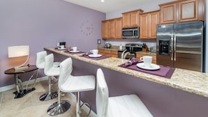 A modern kitchen with a granite countertop, white bar stools, wooden cabinets, stainless steel appliances, and a purple accent wall. - Grhcap8929 - Paradise Palms Resort - 4 Bed 3 Baths Townhouse (Kissimmee)