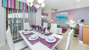 A modern dining area with a white table set for six, purple placemats, and a view of the living room in the background. - Grhcap8929 - Paradise Palms Resort - 4 Bed 3 Baths Townhouse (Kissimmee)