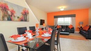 A dining area with a glass table set for four, surrounded by black chairs. There is a sofa with orange cushions in the background. - Grhcup8936 - Paradise Palms Resort - 5 Bed 4 Baths Townhouse (Kissimmee)