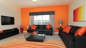 A modern living room with a black sofa, orange cushions, a glass coffee table, and a flat-screen TV. - Grhcup8936 - Paradise Palms Resort - 5 Bed 4 Baths Townhouse (Kissimmee)