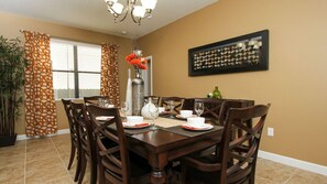 A dining room with a wooden table set for six, surrounded by chairs. There is a vase with flowers on the table, and a framed abstract art piece on the wall.