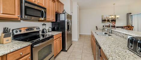 A modern kitchen with wooden cabinets, stainless steel appliances, granite countertops, and a tiled floor.
