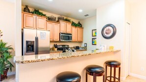 A kitchen with wooden cabinets, a stainless steel refrigerator, a microwave, a gas stove, and a granite countertop. There are two stools and a clock on the wall.