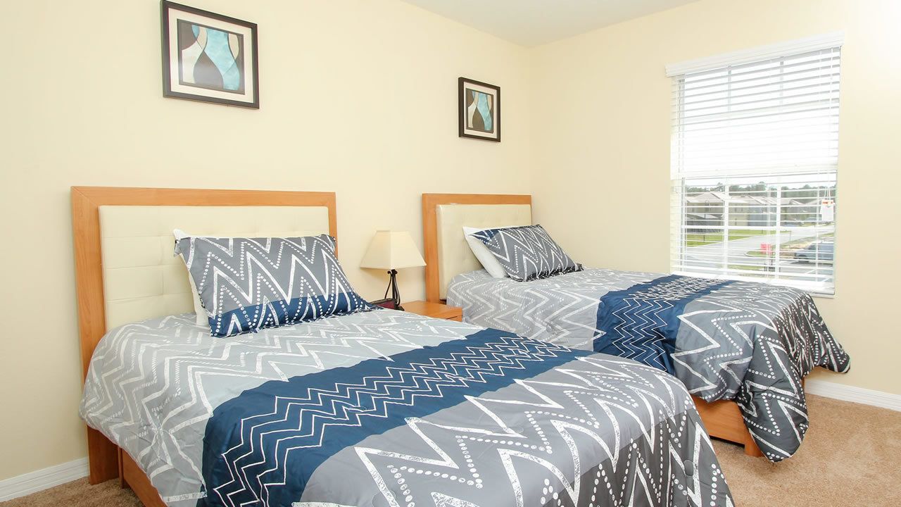 A bedroom with two beds, each with a patterned bedspread and matching pillows. There are two framed pictures on the wall, and a window with blinds.