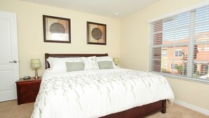 A bedroom with a large bed, two framed pictures on the wall, a nightstand with a lamp, and a window with blinds.
