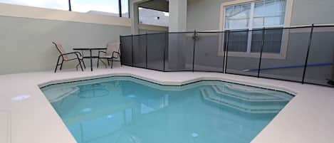 A swimming pool with a glass enclosure, surrounded by a covered patio area with chairs and a table.