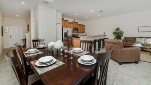 A modern kitchen with wooden cabinets and a central island, a dining area with a wooden table set for four, and a living area with a brown sofa and patterned chairs. - Grhcat8968 - Paradise Palms Resort - 4 Bed 3 Baths Townhouse (Kissimmee)