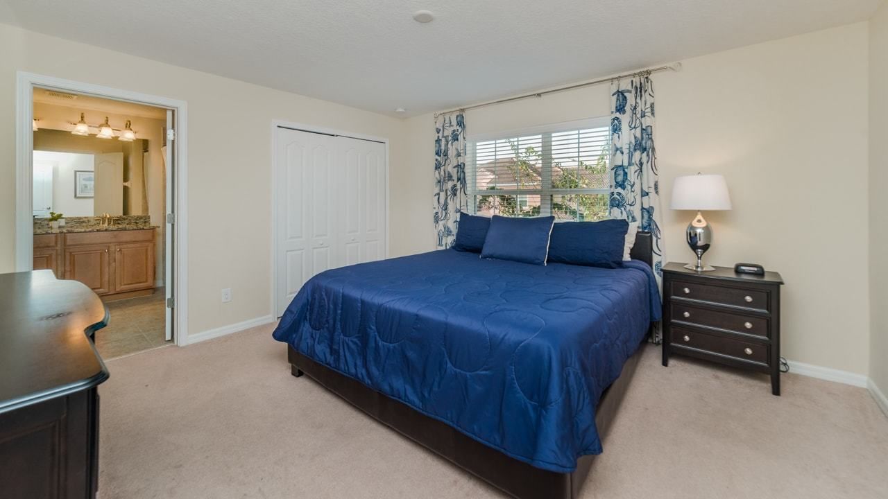 A bedroom with a large bed, a nightstand with a lamp, a dresser, and a door leading to a bathroom.