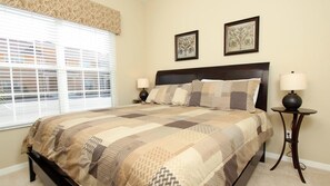 A bedroom with a large bed, two bedside tables with lamps, a window with blinds, and two framed floral artworks on the wall.