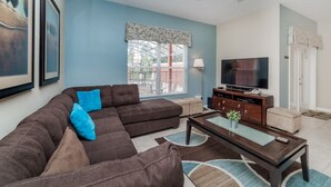 A living room with a sectional sofa, a coffee table, a TV, and a window with blinds. - Grhmap8975 - Paradise Palms Resort - 4 Bed 3 Baths Townhouse (Kissimmee)