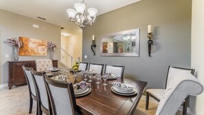 A dining room with a wooden table set for six, surrounded by chairs, a chandelier, and wall-mounted sconces. - Grhbis8974 - Paradise Palms Resort - 5 Bed 5 Baths House (Kissimmee)