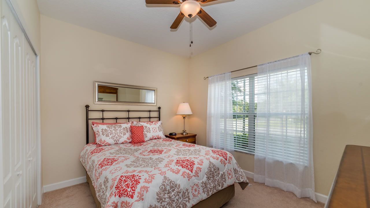 A bedroom with a bed, a nightstand, a lamp, a ceiling fan, a window with blinds, and a mirror on the wall.