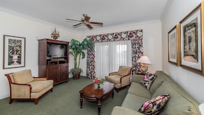 Ein Wohnzimmer mit einer Couch, zwei Sesseln, einem Couchtisch, einer TV-Bank, einem Deckenventilator und einem großen Fenster mit floralen Vorhängen.