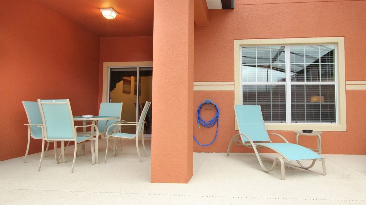 A covered outdoor seating area with four chairs and a table, a blue hose, and a window with blinds.