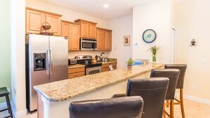 A kitchen with granite countertops, wooden cabinets, a refrigerator, a microwave, a stove, and a sink. There are four bar stools and a potted plant on the counter. - Grhcal8951 - Paradise Palms Resort - 5 Bed 4 Baths Townhouse (Kissimmee)