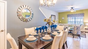 Een eethoek met een donker houten tafel voor vier personen, omringd door witte stoelen. Aan de muur hangt een grote spiegel met een decoratieve lijst, en erboven een kroonluchter.