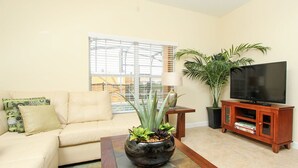 A living room with a beige sofa, a coffee table with a plant, a TV on a wooden stand, and a window with blinds.