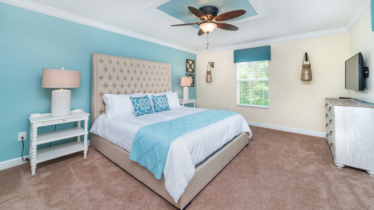 A bedroom with a tufted headboard, a bed with blue and white bedding, a white nightstand with a lamp, a ceiling fan, a TV, and a dresser.